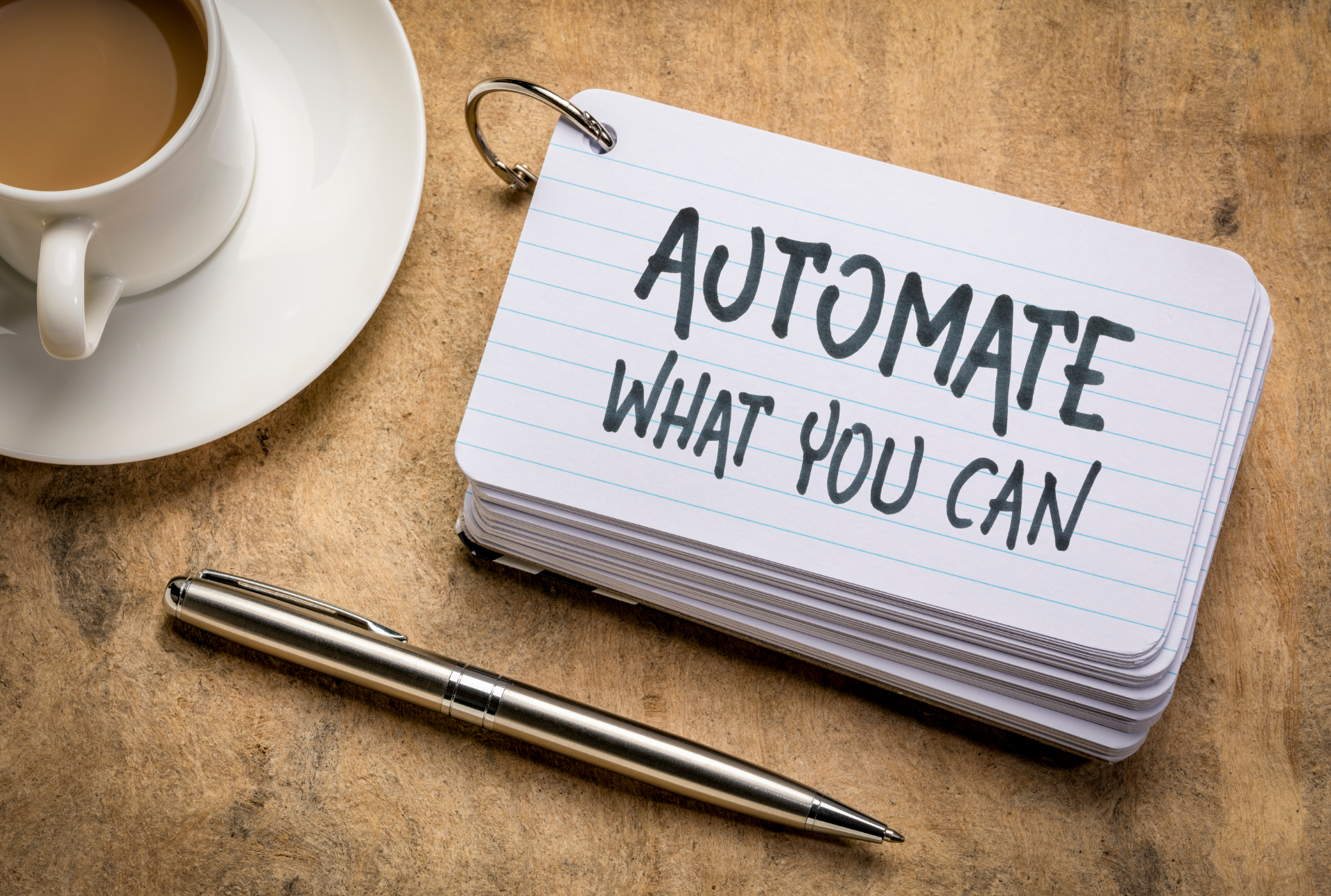 A coffee, pen and paper on a wooden surface with the text 'Automate What You Can' written on it